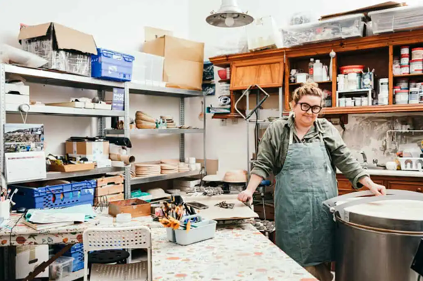 Picture of Ola standing in her studio surrounded by shelves items and equipment.